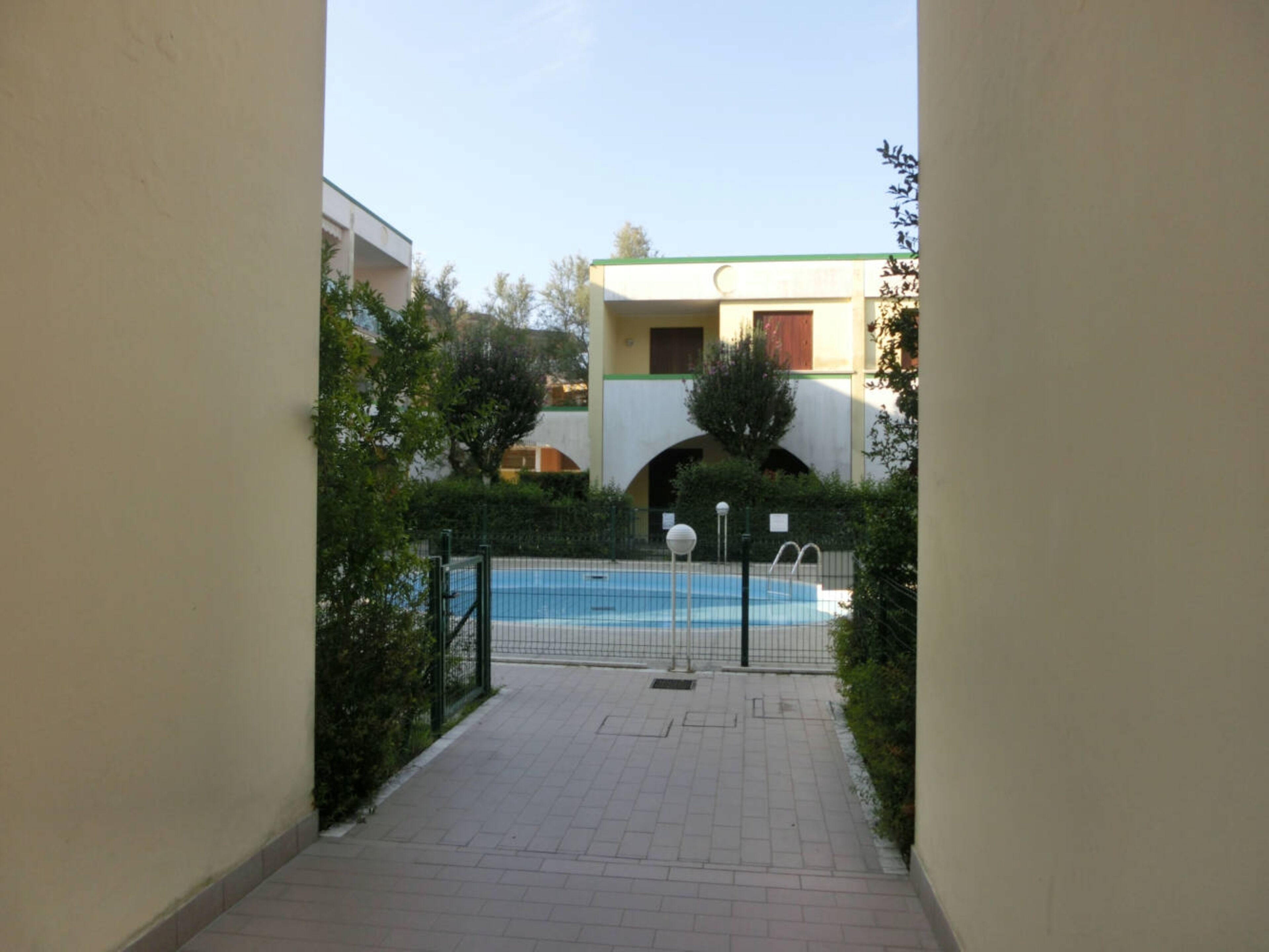 A pathway leading to a swimming pool with a fence, surrounded by greenery and buildings in the background.