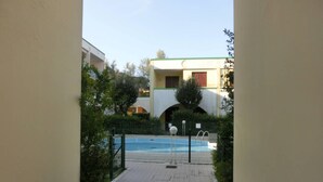 A pathway leading to a swimming pool with a fence, surrounded by greenery and buildings in the background.