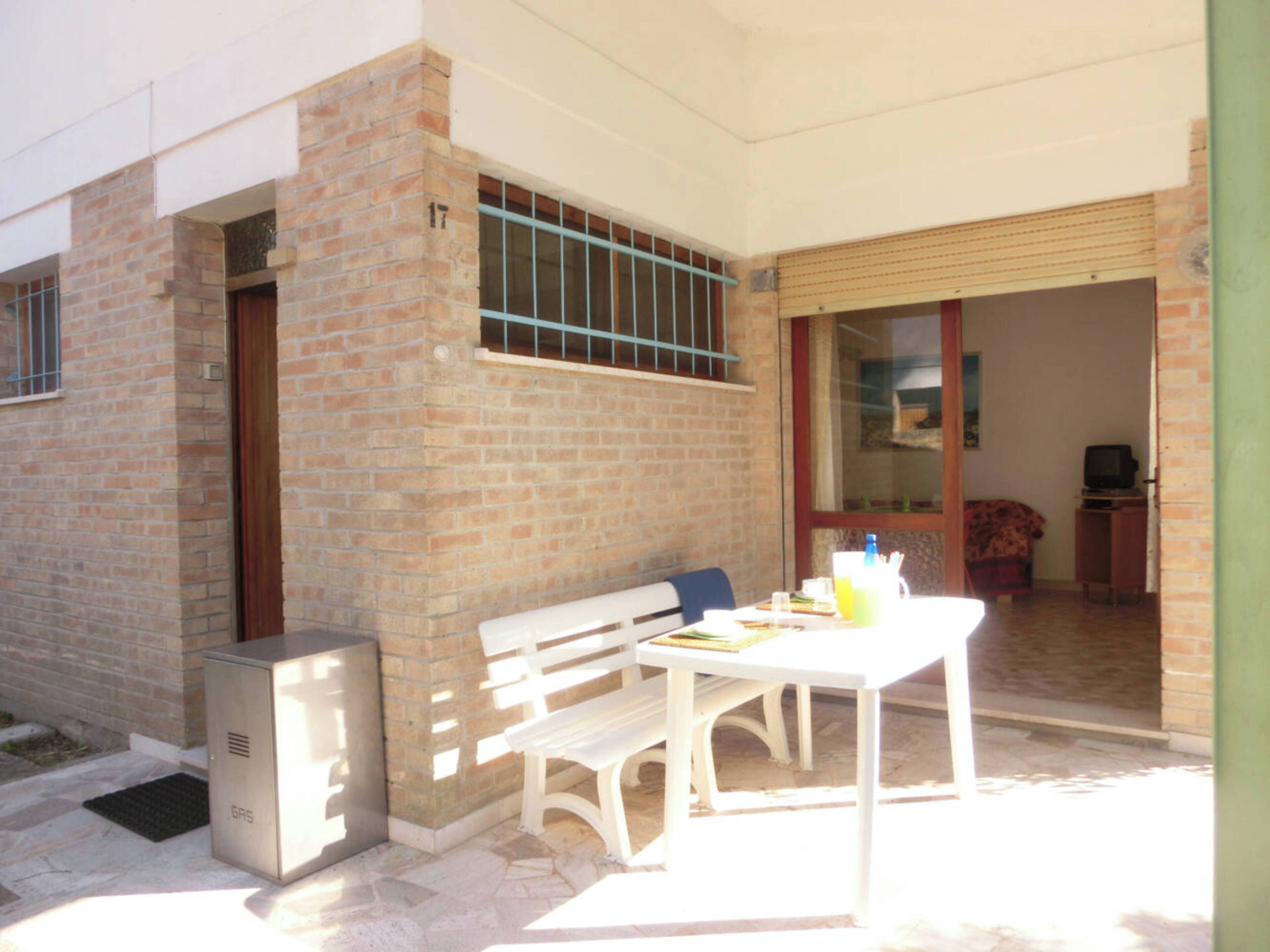 An outdoor patio with a white table and bench, a small refrigerator, and a sliding glass door leading to an interior room.