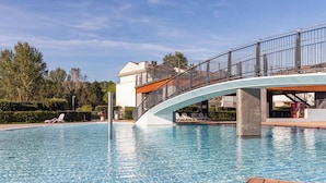 A swimming pool with a wooden deck in the foreground, a bridge with railings, and lounge chairs along the poolside. - Wonderful Flat With Solarium and Swimming Pools (San Michele al Tagliamento)