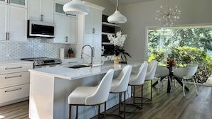 A modern kitchen with white cabinetry, a central island with bar stools, pendant lighting, and a view of greenery outside.