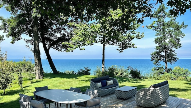 An outdoor deck with a round table and chairs, a sofa, and a cushioned bench, overlooking a body of water.