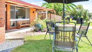 Une terrasse avec une table et des chaises, un grand parasol, et une maison en briques dotée d’un porche grillagé.