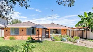 Une maison à deux étages, avec un toit en tuiles, une terrasse couverte et un petit jardin à l’arrière, comprenant une pelouse et des plantes en pot.