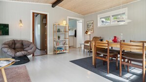 En rummelig opholdsstue med højt til loftet, et spisebord med stole, en bogreol og en behagelig lænestol.