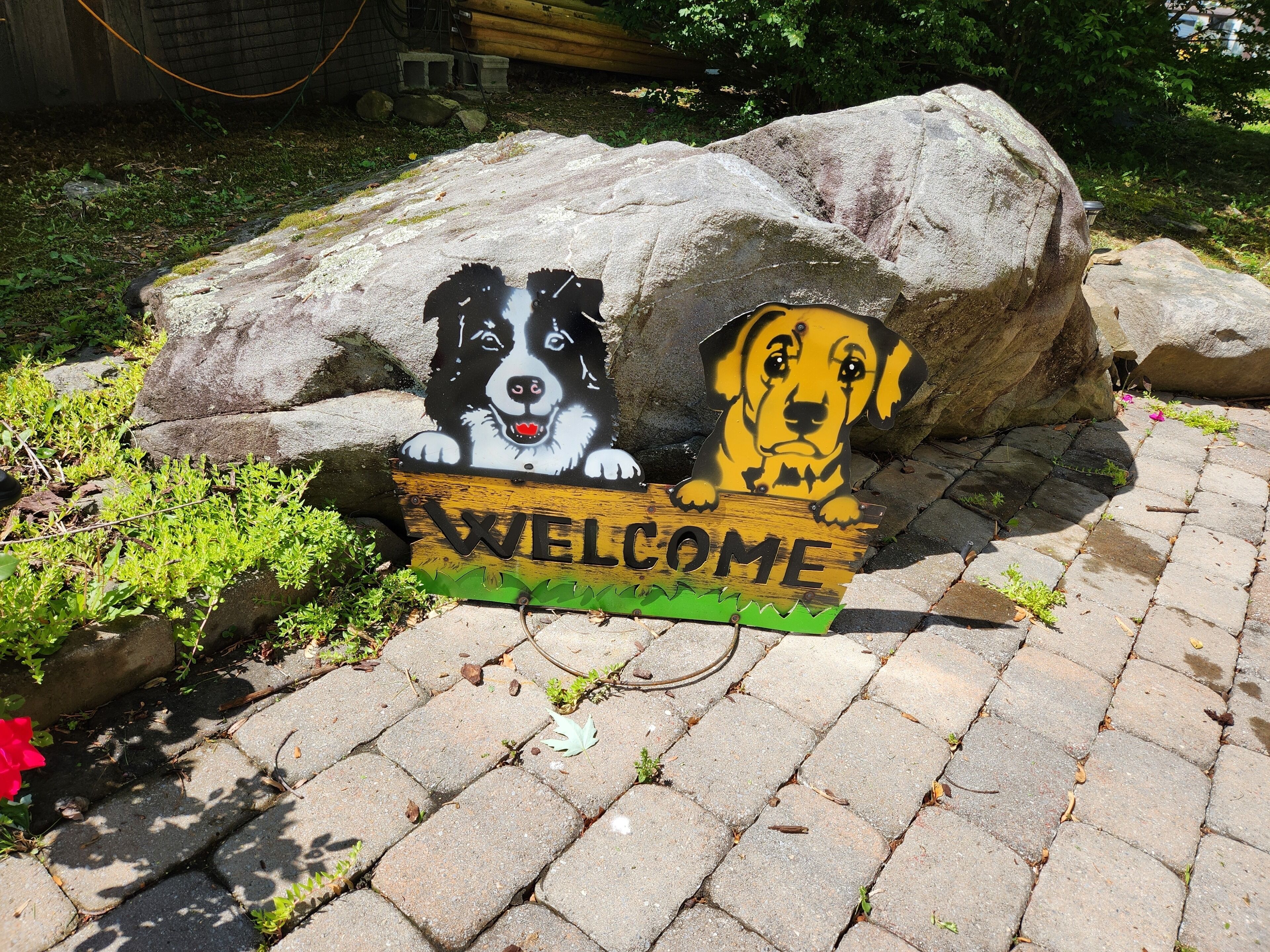 A welcome sign with two dog faces, a large rock, and a paved area with greenery.