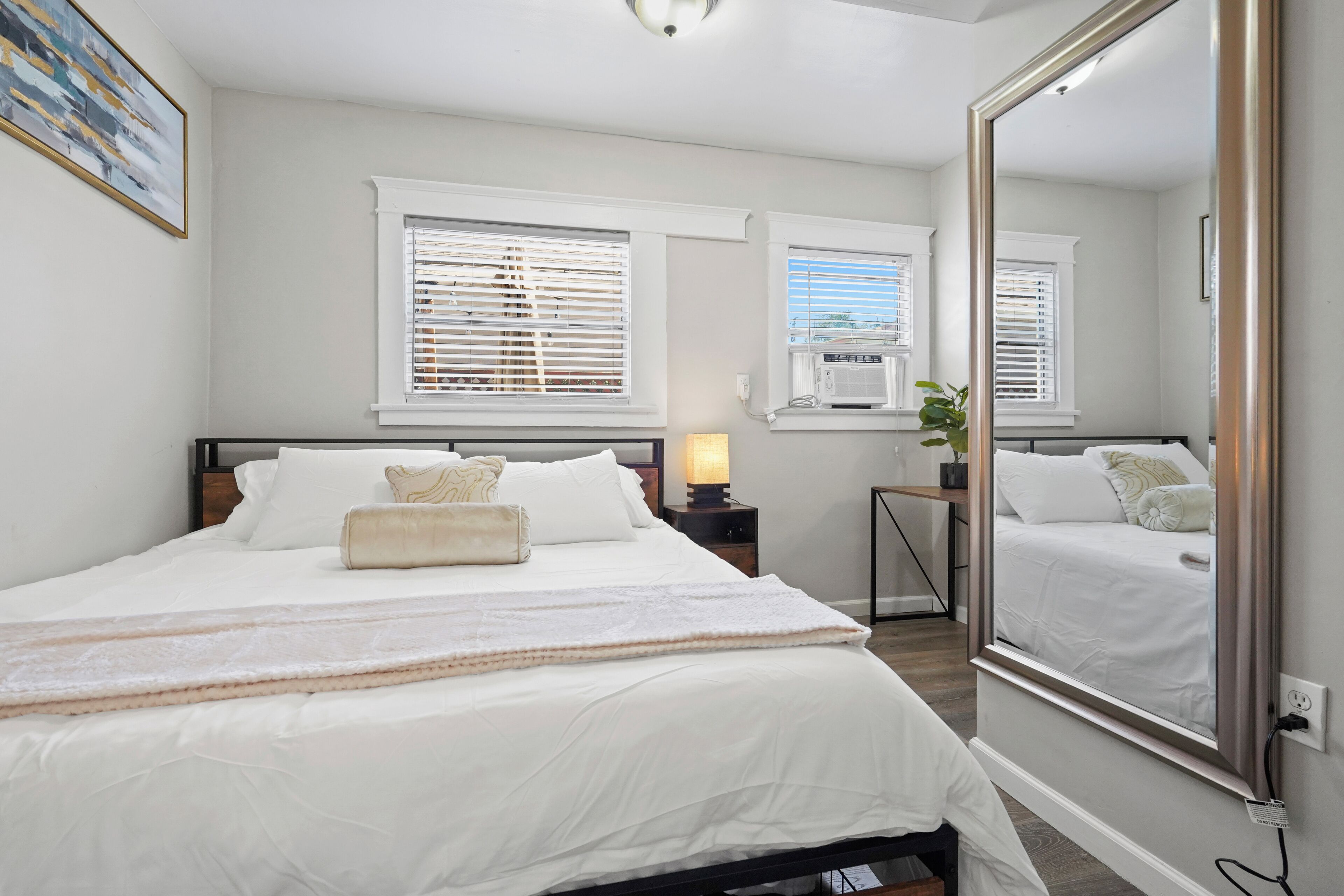 A bedroom with a large bed, two windows with blinds, a bedside table with a lamp, and a large mirror.