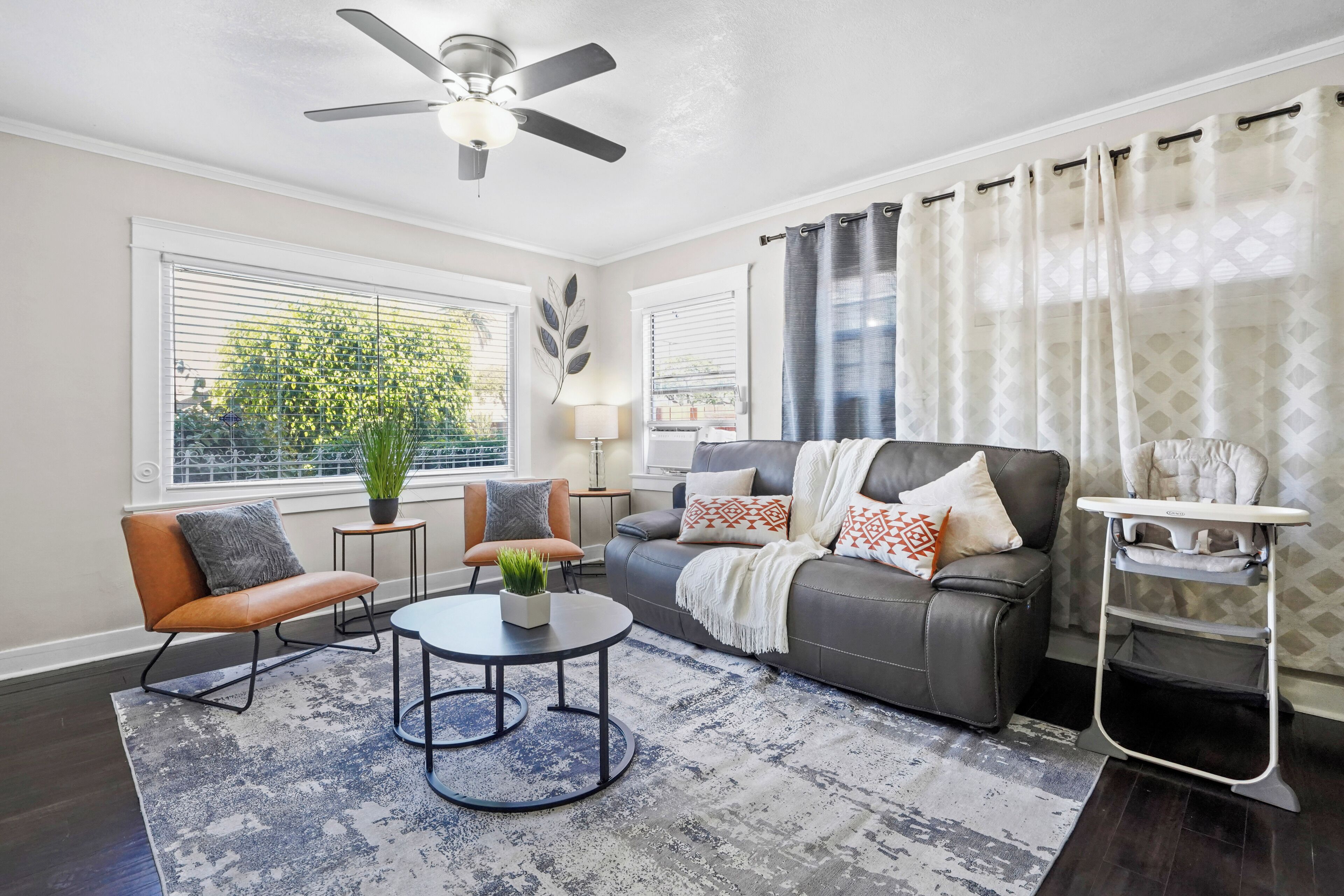 A modern living room with a grey sofa, a round coffee table, a ceiling fan, and a view of a tree through the window.