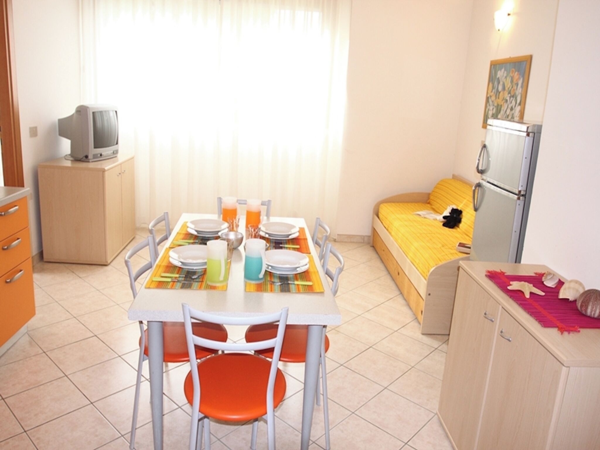 Habitación pequeña con una mesa de comedor para dos, una cama con una manta amarilla, una kitchenette y un televisor sobre un mueble.