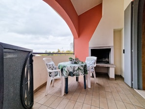 A balcony with a grill, two chairs, a table with a leaf-patterned tablecloth, and a view of the sky. - Bright Apartment With Swimming Pool (San Michele al Tagliamento)