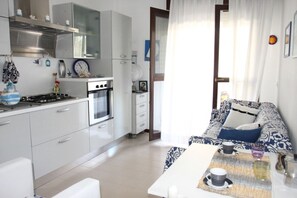Una cocina moderna con armarios blancos, una estufa y un horno. Hay una zona de comedor con una mesa y sillas, y un sofá con cojines estampados.