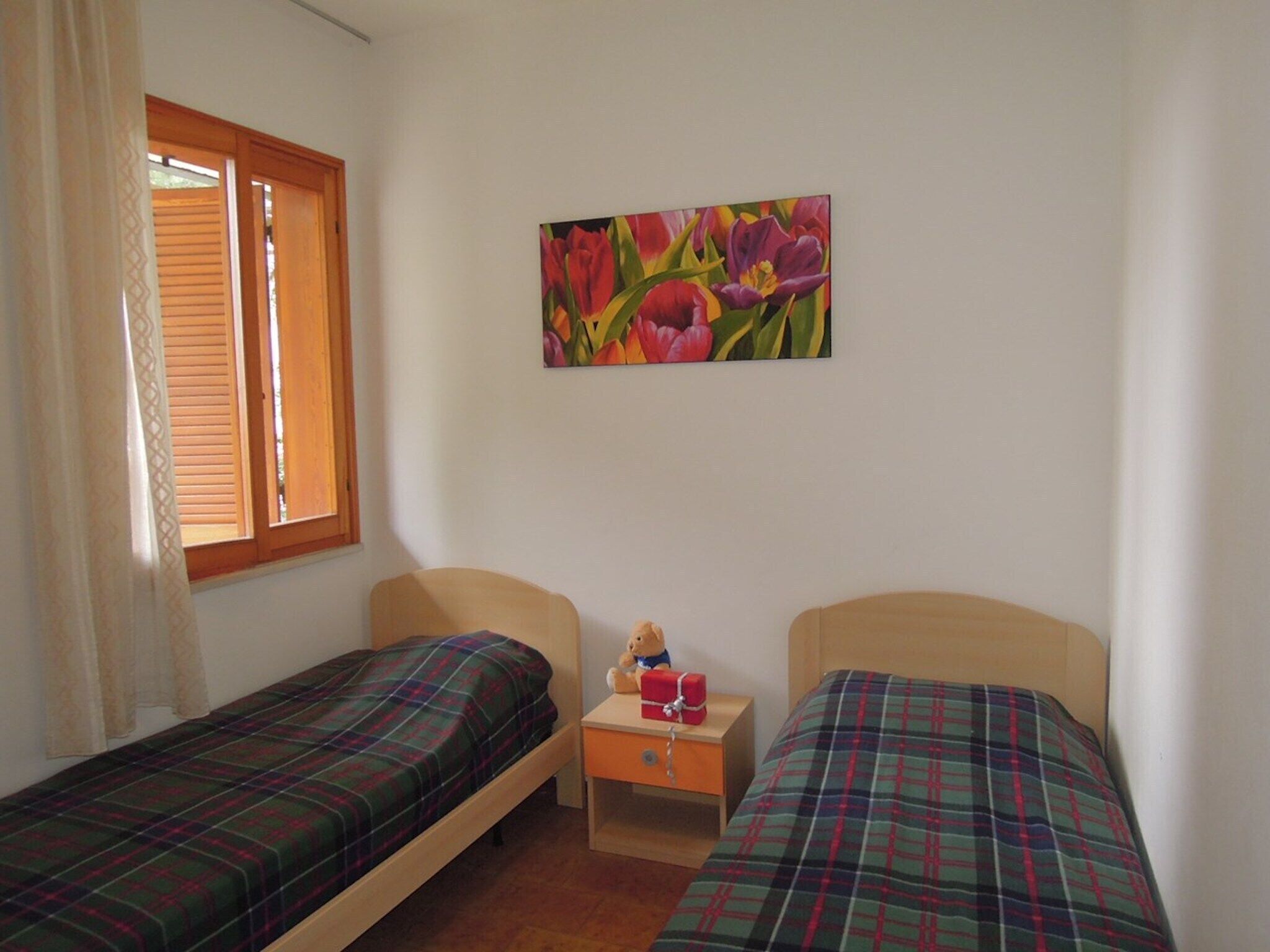 A room with two single beds, a small bedside table with a teddy bear, a window with wooden blinds, and a floral painting on the wall.