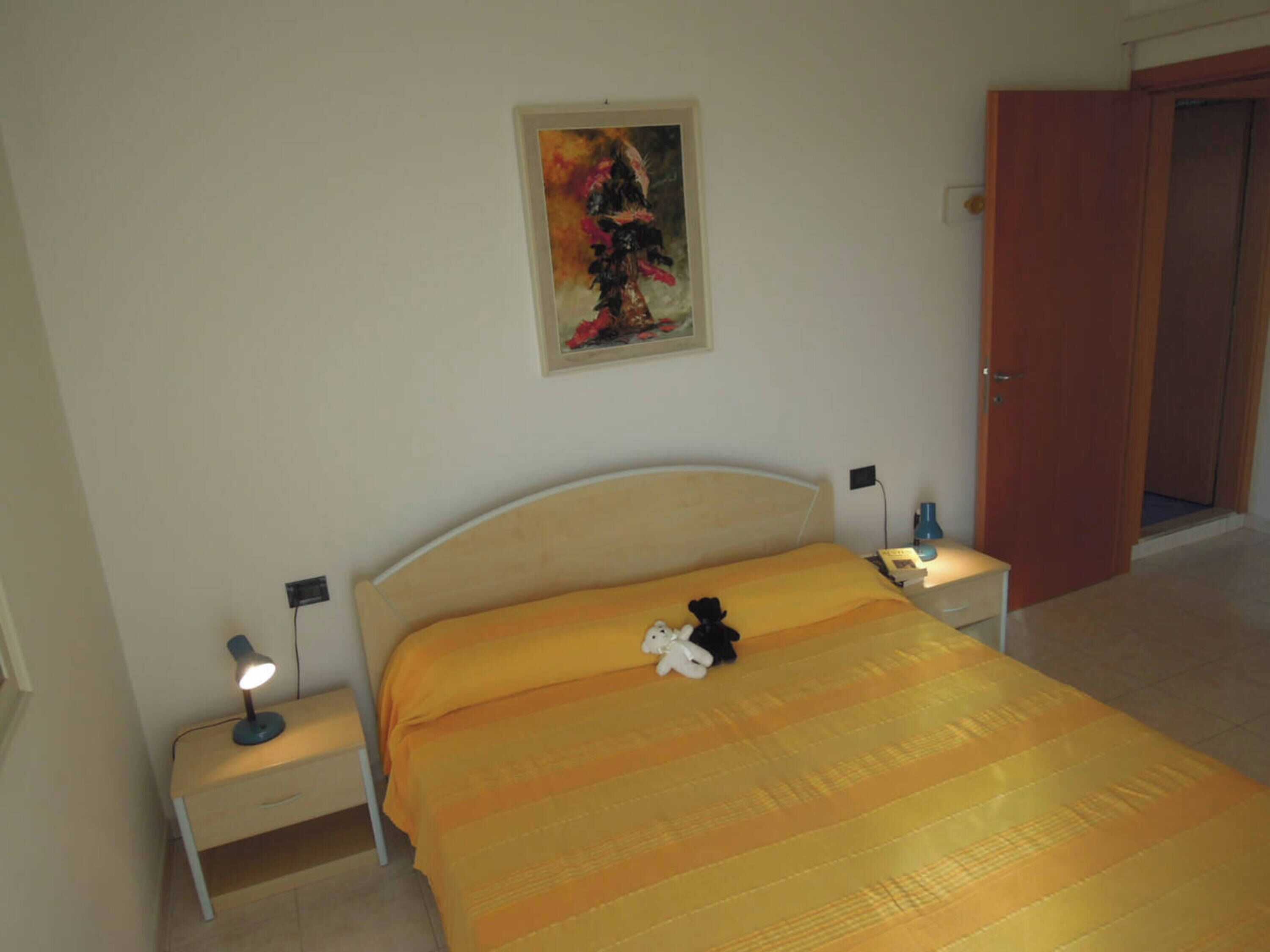 A bedroom with a bed, bedside tables, a nightstand, a lamp, a teddy bear, a painting on the wall, and a door.