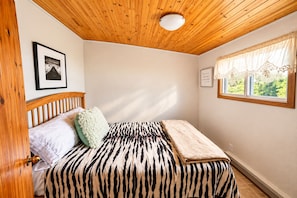 A bedroom with a wooden ceiling, a bed with a zebra-striped bedspread, a green pillow, and a window with a view of greenery.