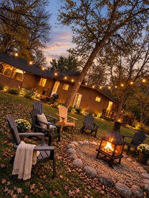 Un espace extérieur accueillant, aménagé avec des fauteuils Adirondack et un foyer, entouré d’un chemin de pierres et d’un bâtiment en bois illuminé de guirlandes lumineuses.