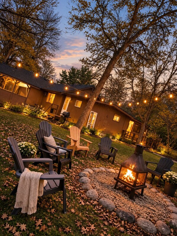 Cómoda zona de estar al aire libre con sillas Adirondack y un hogar para fuego, rodeada por un sendero de piedra y un edificio de madera con luces colgantes.