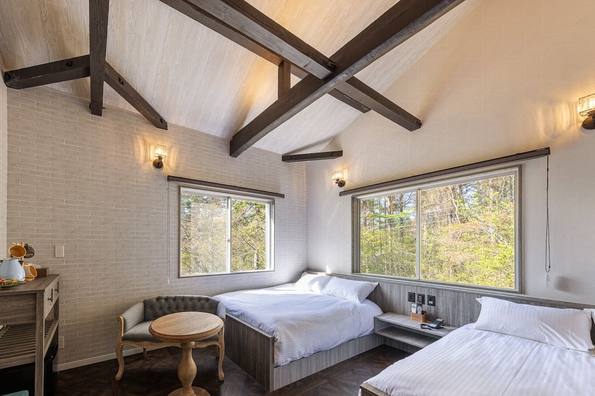 A bedroom with two beds, a small table, and a chair. There are windows with views of trees.