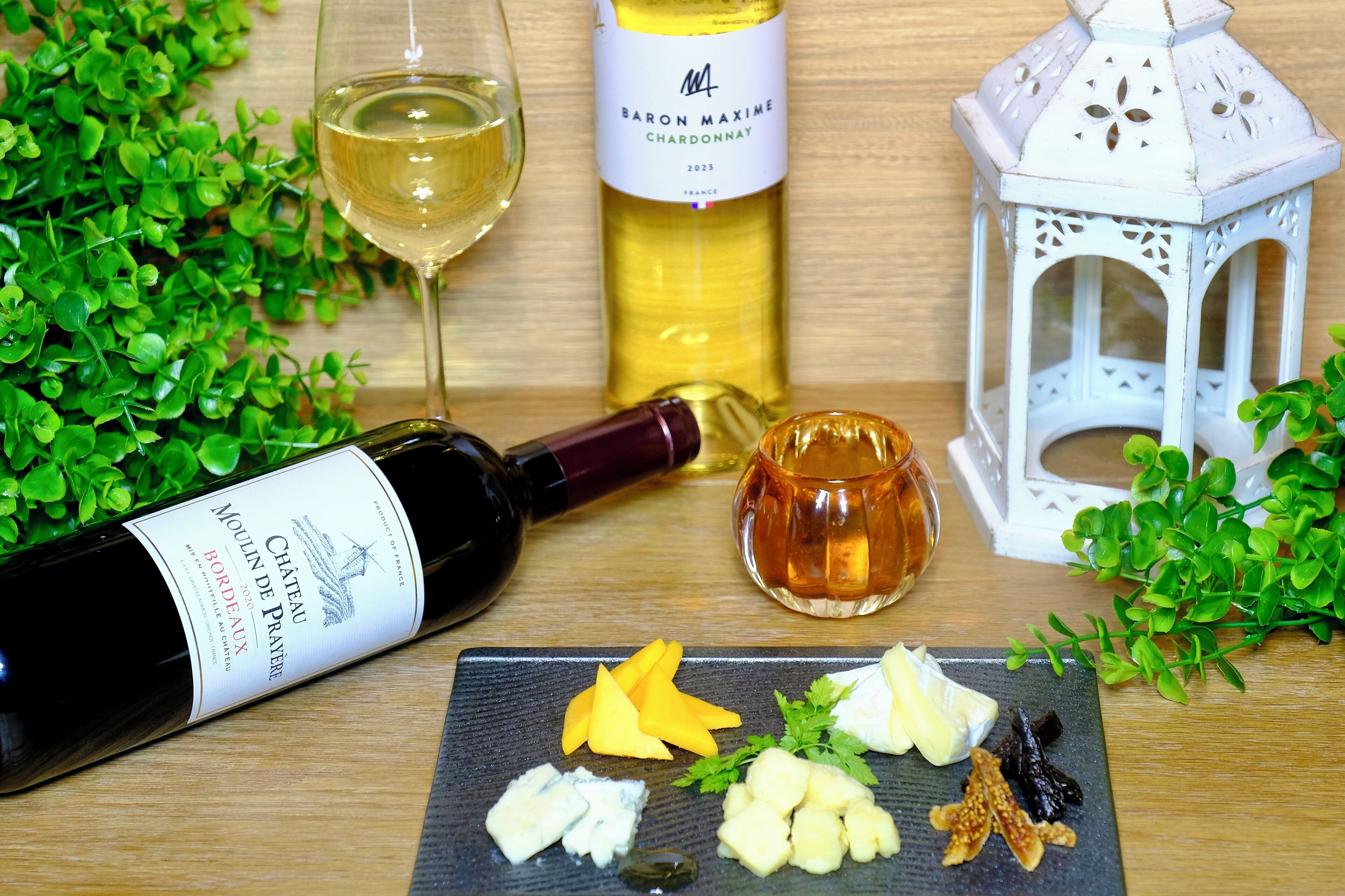 Eine Flasche Chateau de Pomerol 2015 Bordeaux Wein, ein Glas Weißwein, eine Flasche Baron Maxime Chardonnay 2023, eine kleine Schale Honig und ein weißer Laternen auf einem Holztisch mit Grünpflanzen.