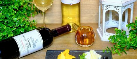Eine Flasche Chateau de Pomerol 2015 Bordeaux Wein, ein Glas Weißwein, eine Flasche Baron Maxime Chardonnay 2023, eine kleine Schale Honig und ein weißer Laternen auf einem Holztisch mit Grünpflanzen.