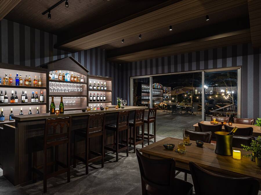 Un espace bar doté de comptoirs et de tabourets en bois, d’étagères remplies de bouteilles et de verres, et offrant une vue sur l’espace extérieur aménagé pour les clients.
