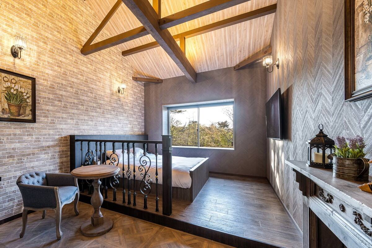A cozy room with a brick wall, wooden ceiling beams, a bed, a small table with chairs, and a window with a view.