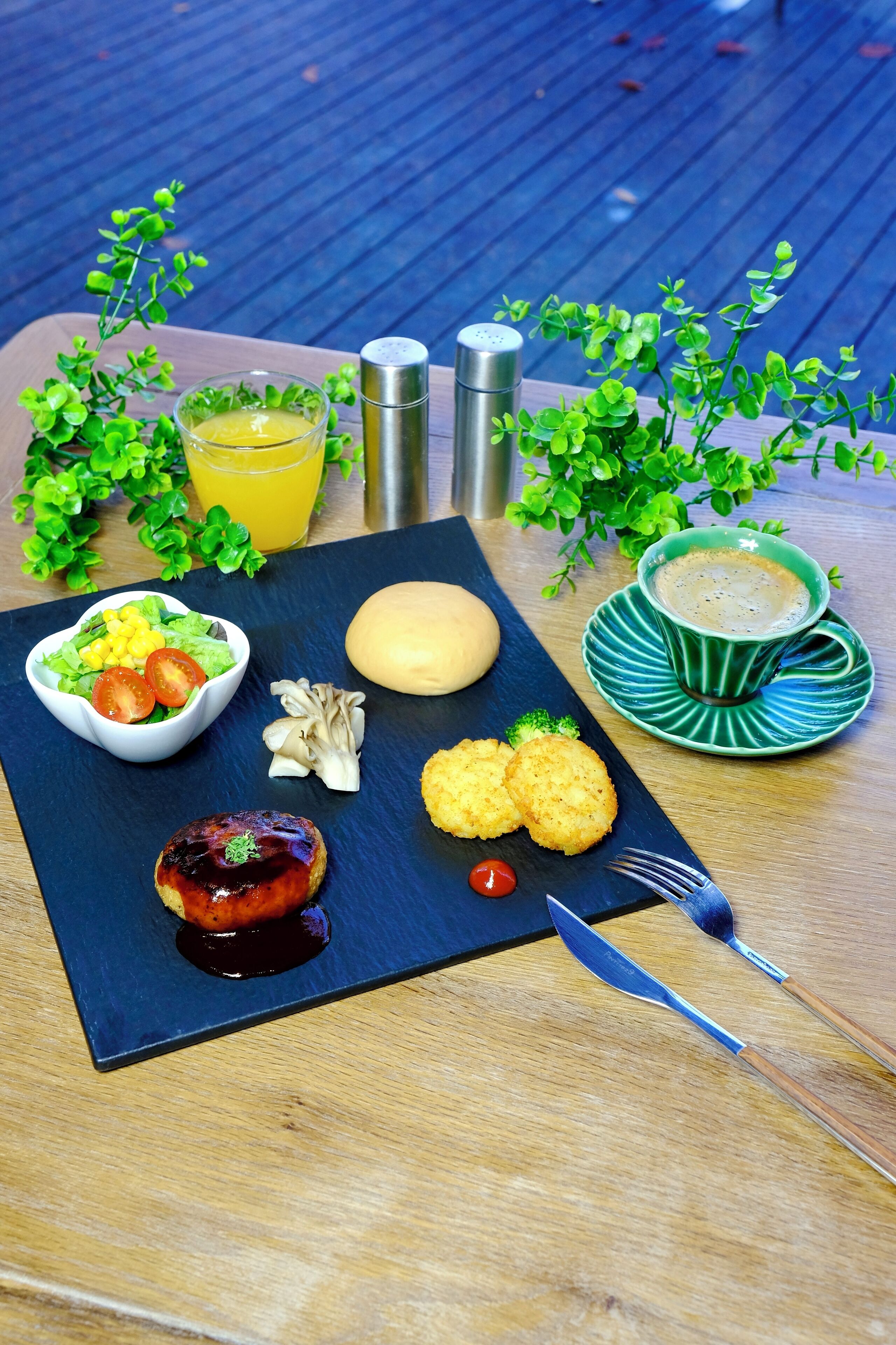 A meal with a salad, grilled meat, and bread on a slate board, accompanied by a cup of coffee and a glass of orange juice.