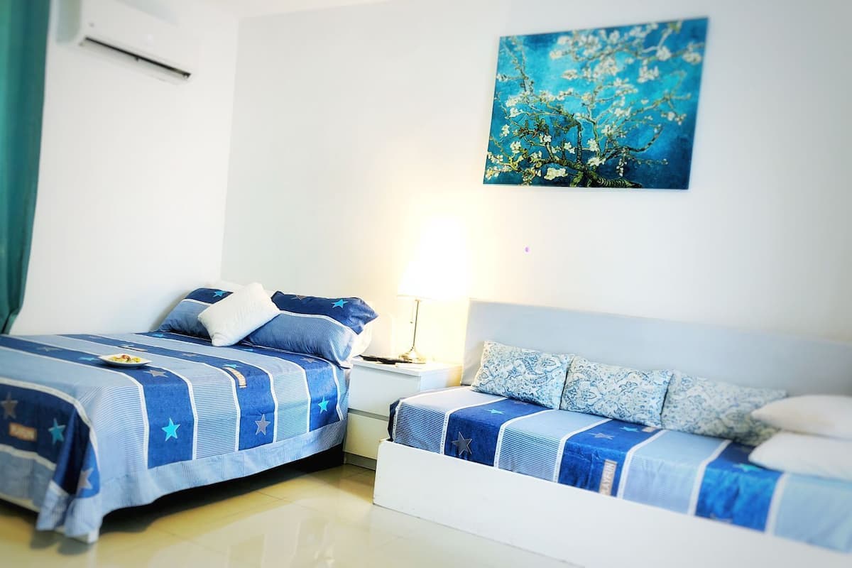 A hotel room with two beds, each with a blue and white striped bedspread and pillows, a small bedside table with a lamp, and a wall-mounted painting of a tree.