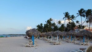 A beach with rows of sun loungers and thatched umbrellas, palm trees, and a clear sky. - Punta Cana (Punta Cana)