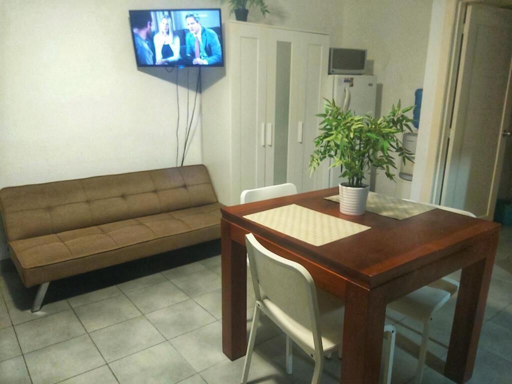 Ein Wohnzimmer mit einer Couch, einem Holzesstisch mit einem Stuhl, einem an der Wand montierten Fernseher und einem Blumentopf.