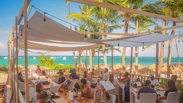 An outdoor dining area with tables and chairs, overlooking a beach with palm trees and the ocean. - Punta Cana (Punta Cana)