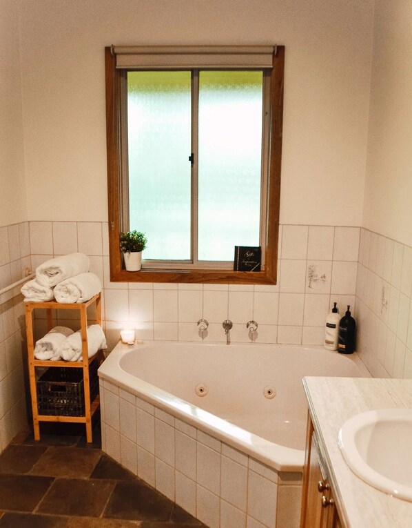 A bathroom with a large bathtub, a window with a view, a shelf with towels, and toiletries. - Shaw Thing Lodge: Wifi, Foxtel, Pet Friendly. (Eildon)