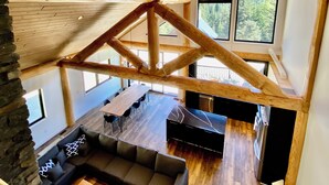 Un vaste espace de vie aménagé dans un style industriel, avec un plafond haut, des poutres apparentes en bois et de grandes fenêtres offrant une vue sur la forêt.