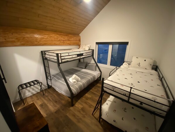 A dormitory room with two bunk beds, a small chair, a window, and a wooden ceiling.