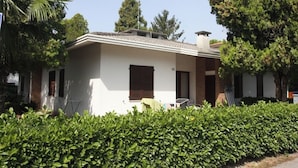 A single-story house with a white exterior, a tiled roof, and a chimney. There is a covered patio area with a table and chairs. - Lovely Villa Just 500mt From the sea (San Michele al Tagliamento)