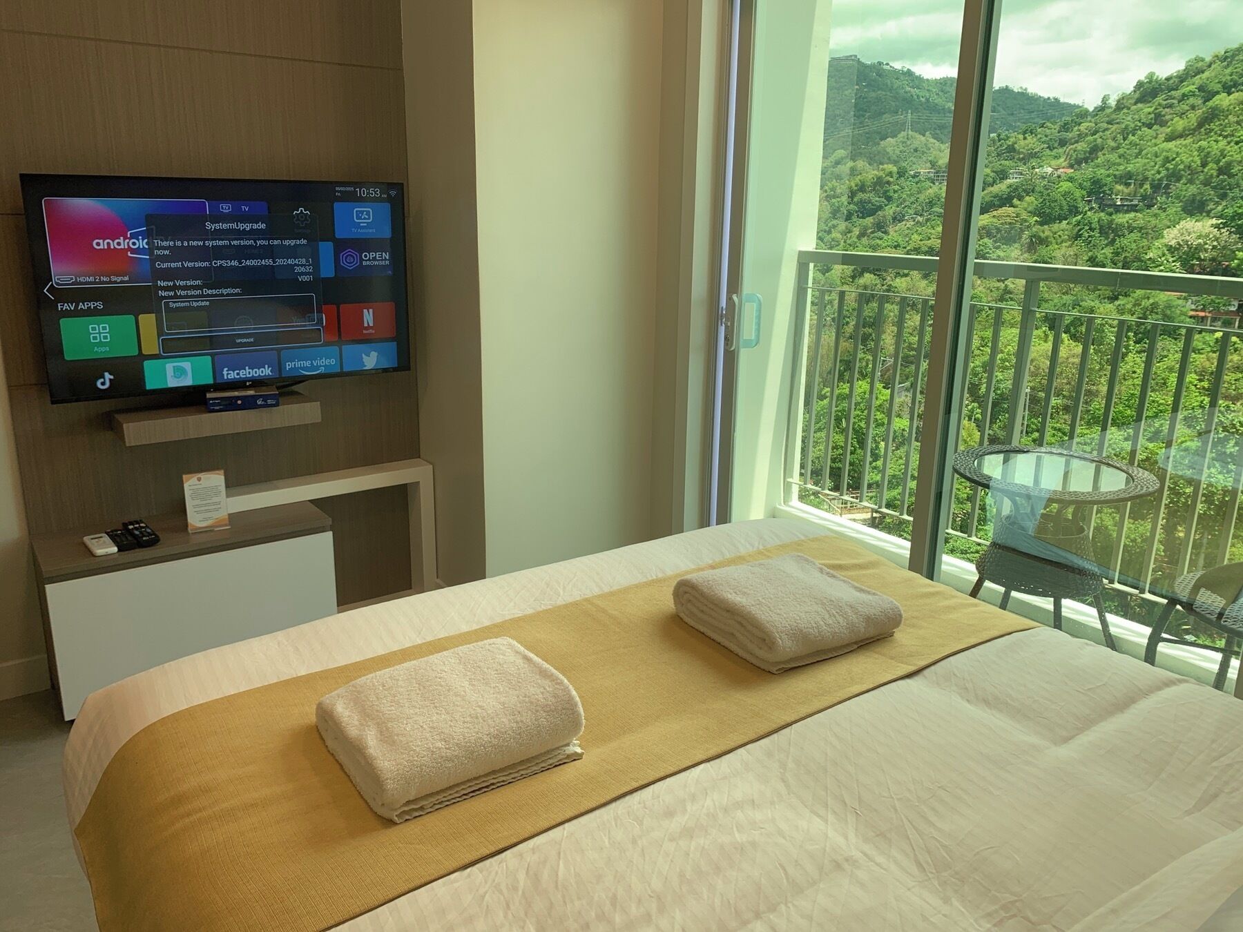A hotel room with a bed, a TV on a stand, a balcony with a view of greenery, and a table with a chair.