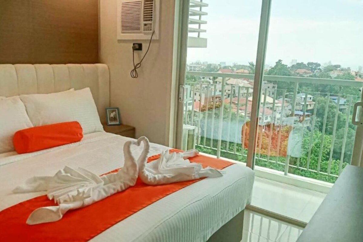 A hotel room with a bed, a window with a view of buildings, and a wall-mounted air conditioner.