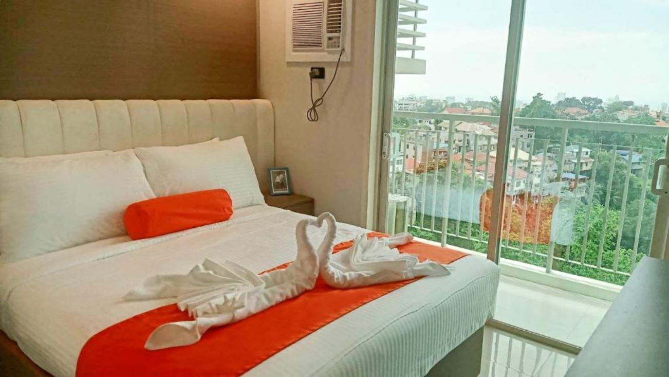 A hotel room with a bed, a window with a view of buildings, and a wall-mounted air conditioner.