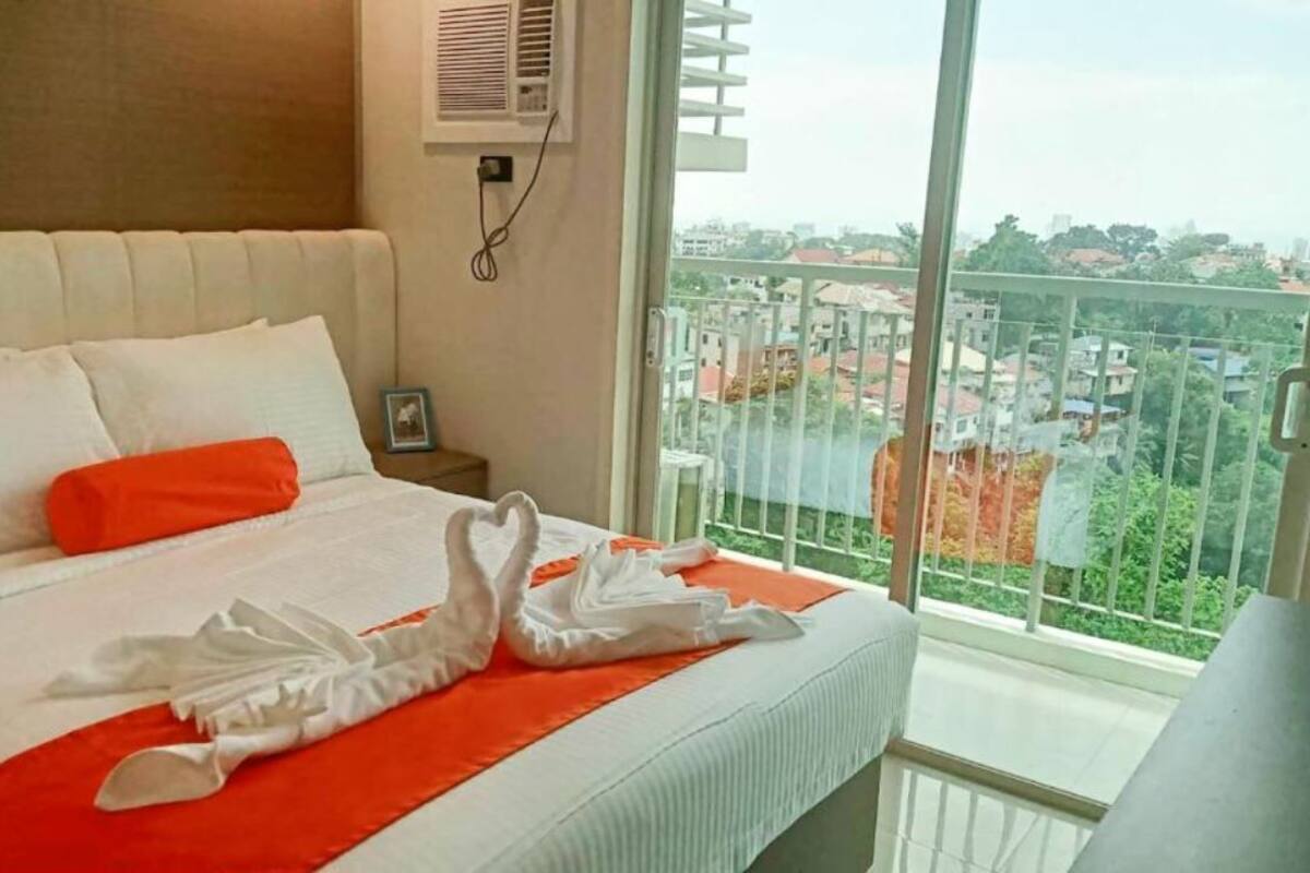 A hotel room with a bed, a window with a view of buildings, and a wall-mounted air conditioner.