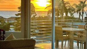 Una habitación con vista al atardecer sobre el océano, un balcón con mobiliario de mimbre y un comedor.