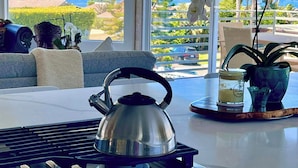 Una cocina con una tetera sobre una estufa a gas, una ventana con vista al océano y una mesa con una planta y una vela.