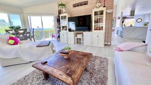 Una sala de estar moderna con un televisor de pantalla plana grande, sofás blancos, una mesa de centro de madera y vistas al exterior.