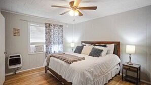 A bedroom with a bed, a ceiling fan, a window with curtains, a bedside table with a lamp, and a wall-mounted heater. - Welcome to Catfish Cabin! (Henderson)