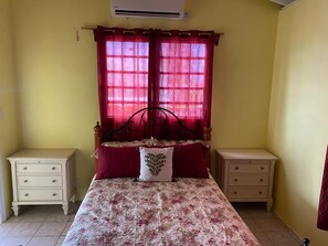 Ein Schlafzimmer mit einem Bett, zwei Nachttischen, einem Fenster mit roten Vorhängen und einer Klimaanlage.