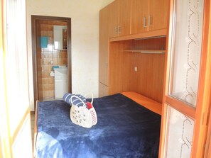 Quarto pequeno e compacto com uma cama, um guarda-roupa de madeira e um banheiro visível através de uma porta aberta.