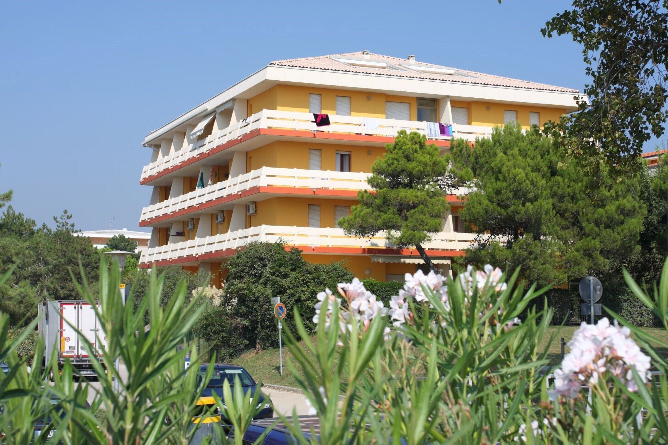 A multi-story yellow building with balconies, surrounded by greenery and parked cars.