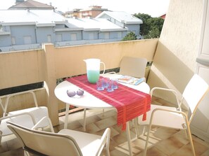 Um pequeno pátio externo com uma mesa redonda, cadeiras brancas, toalha de mesa vermelha e um jarro.