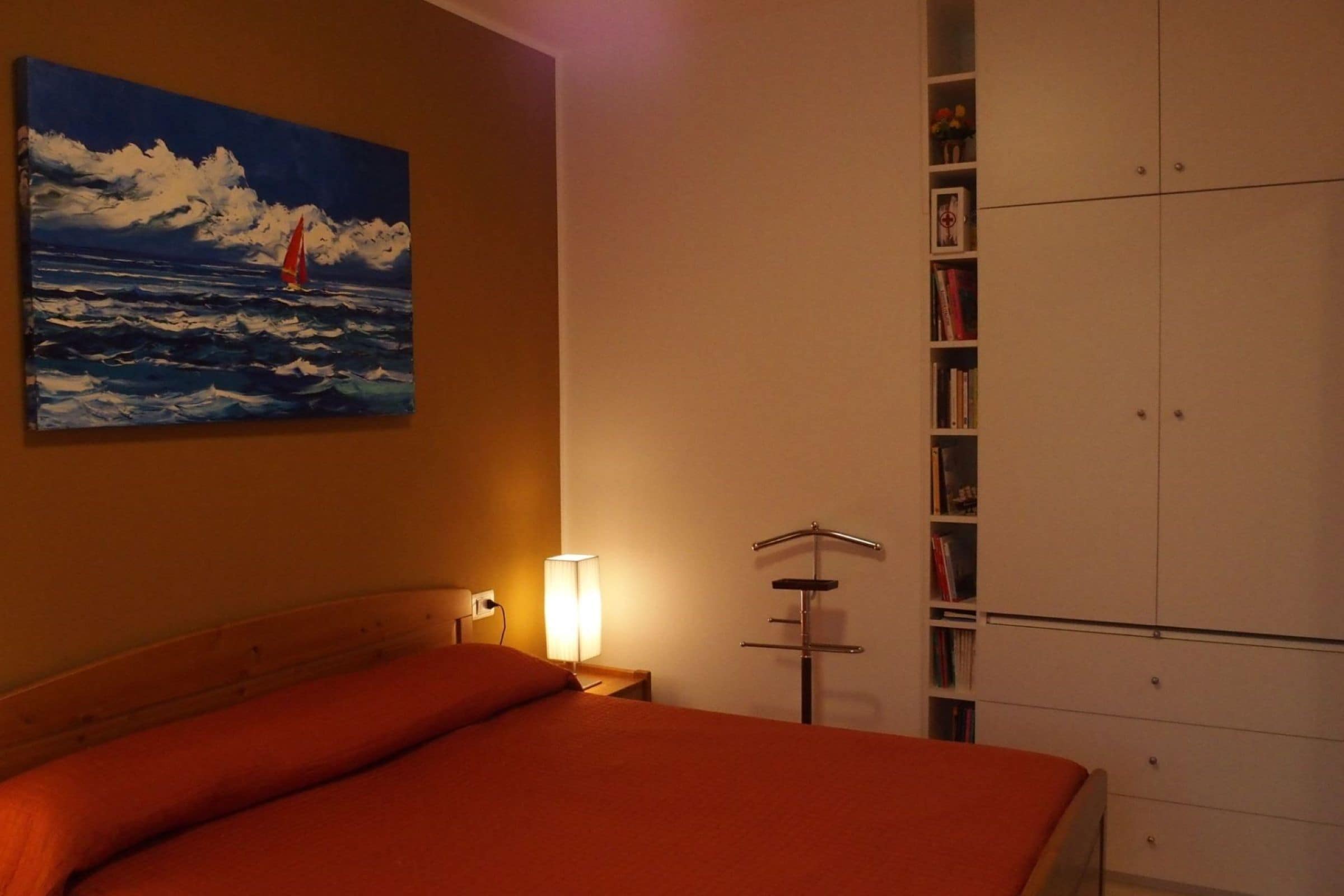 A bedroom with a bed, a painting of a sailboat, a bedside lamp, and a wardrobe.