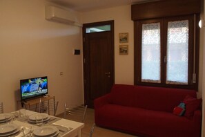 Uma sala de estar com um sofá vermelho, uma televisão sobre um móvel, uma mesa de jantar posta com pratos e copos, e uma porta com painéis de vidro jateado.