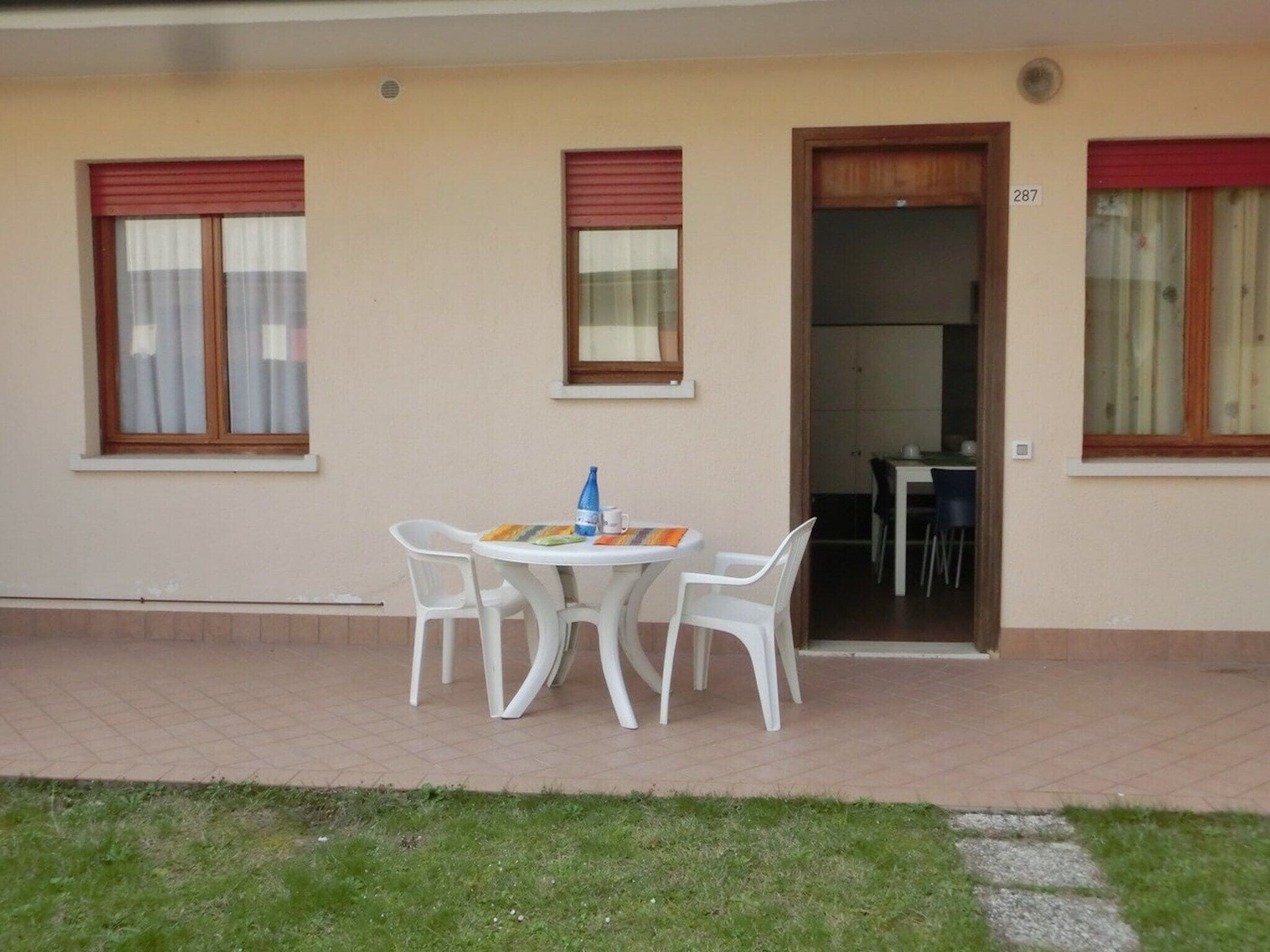 A building with a patio area featuring a table and four chairs. There are two windows with red shutters, one door, and a visible room number 287.