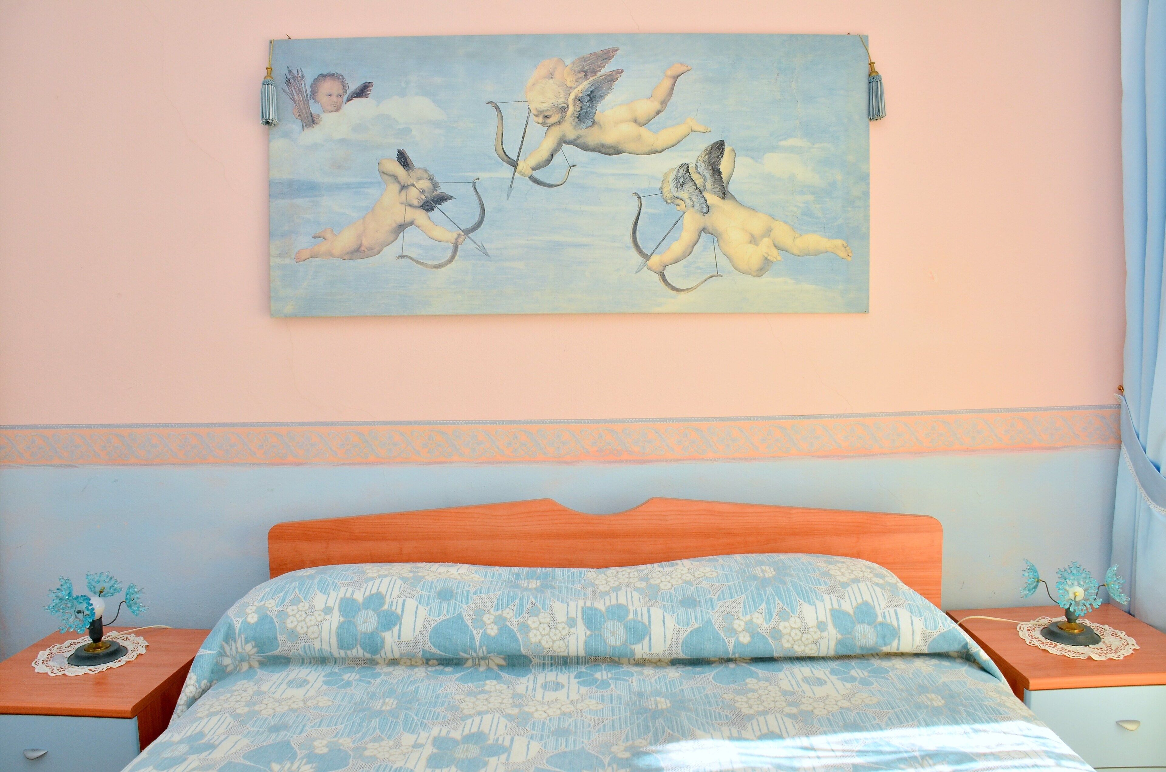 A bedroom with a bed, a nightstand, a bedside table, and a wall painting of cherubs.
